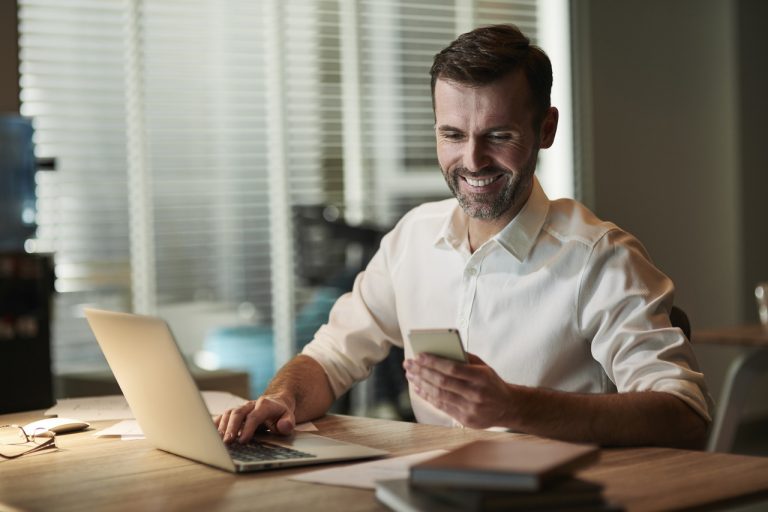 Funcionário de uma empresa usando uma camisa bege com botões, sentado em uma mesa de trabalho diante de um notebook, acessando descontos e promoções de um clube de vantagens White Label no celular